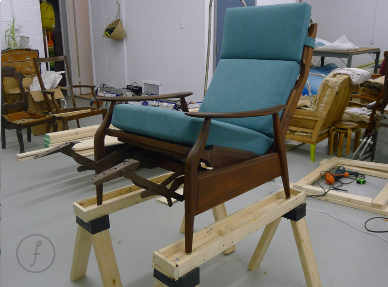 Wooden reclining armchair with teal cushions, raised on two sawhorses in a workshop